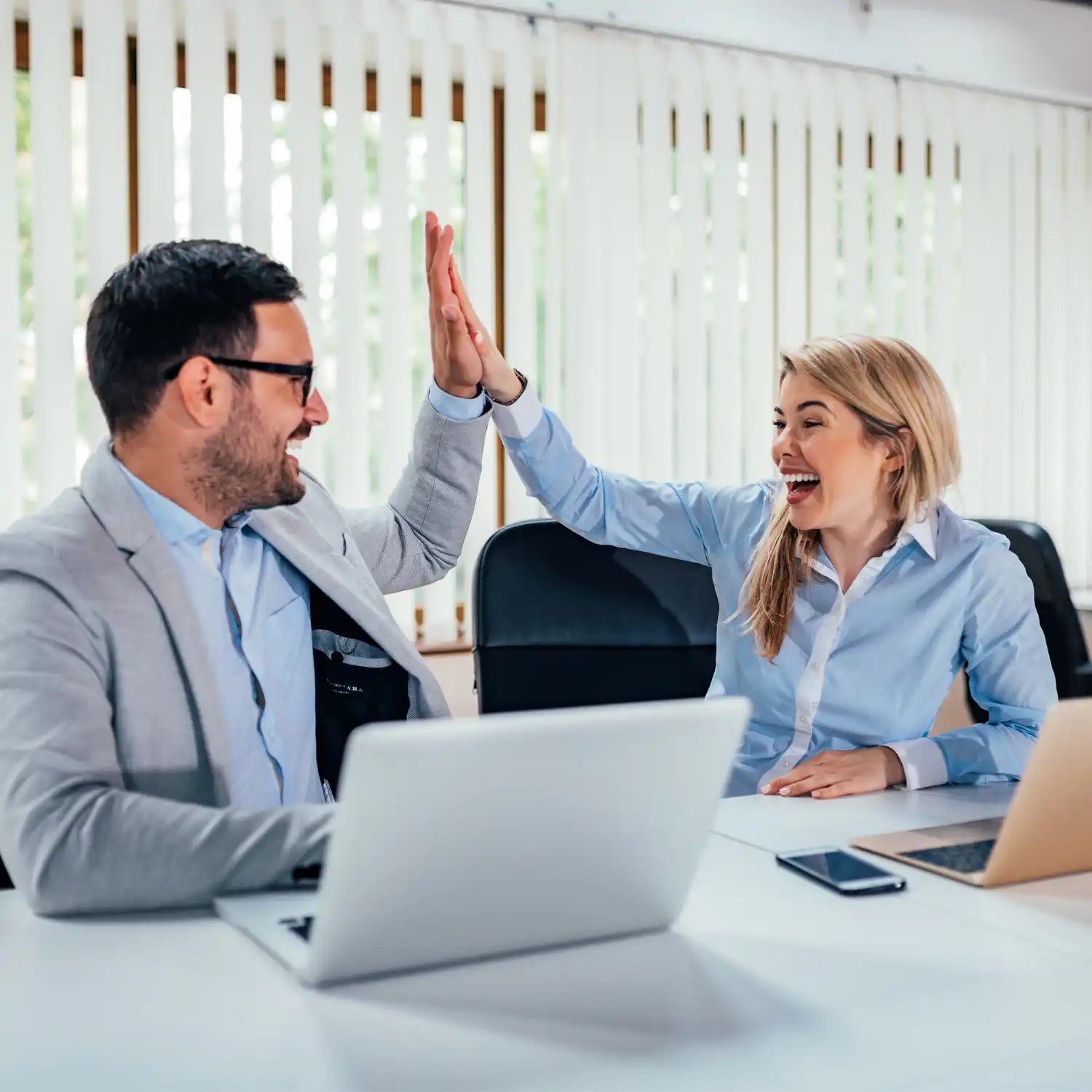 Kaksi työkaveria juhlii innokkaalla viitosella työpöydÀllÀÀn.