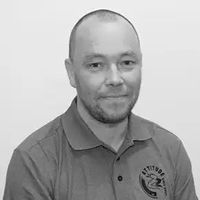 Musta-valkoinen lÀhipotretit kuva henkilöstÀ, joka pystyy polo-paidassa, jossa on pyöreÀ logo.