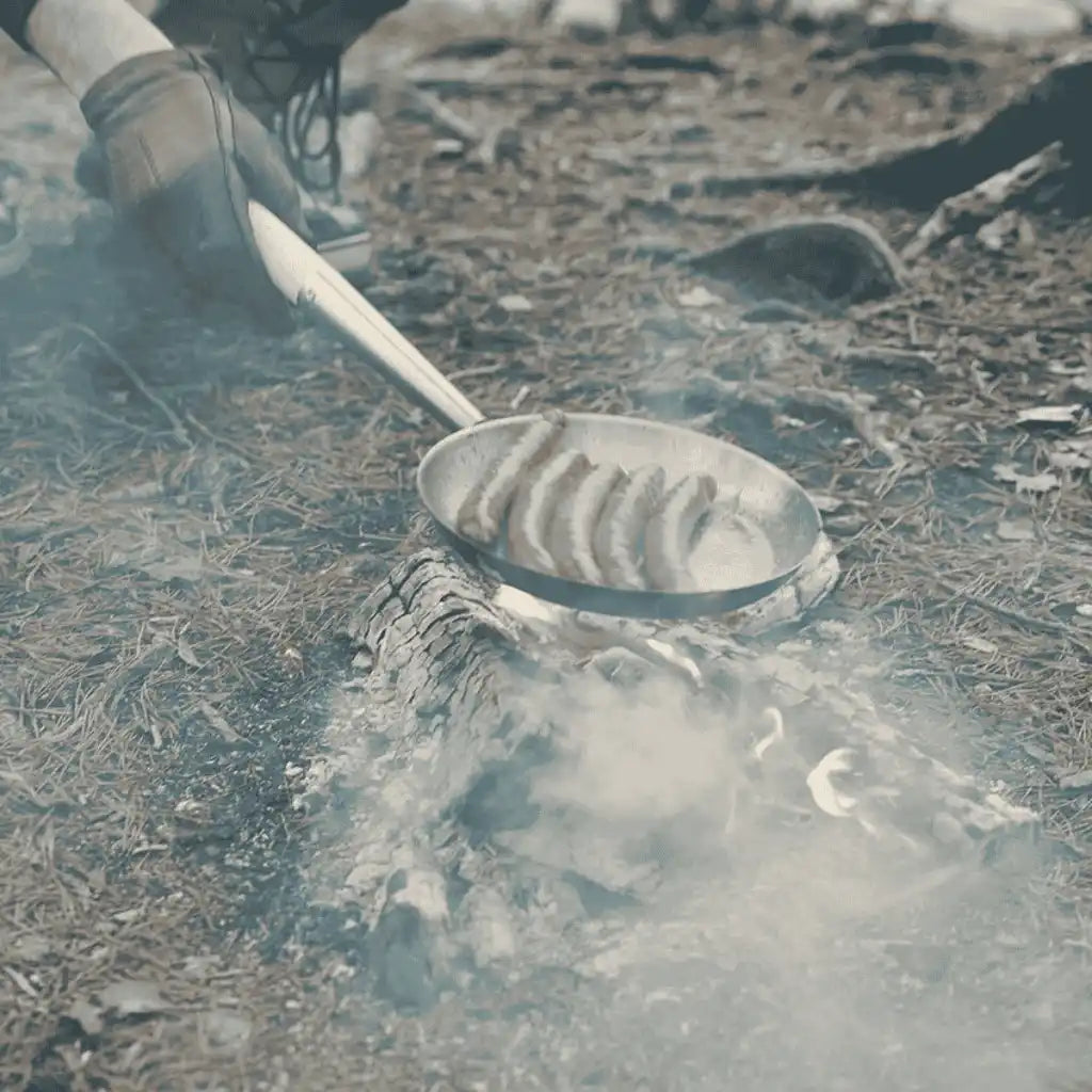 Mies paistaa makkaraa nuotiolla Vinga Vici eräpannun pitkällä kahvalla – turvallinen henkilöstölahja